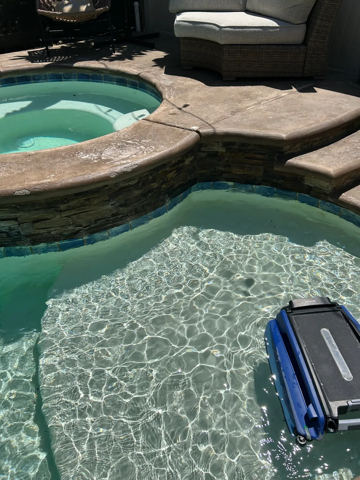 Aerial view of a robotic pool cleaner operating in a freeform pool with attached round spa during professional pool and spa cleaning service
