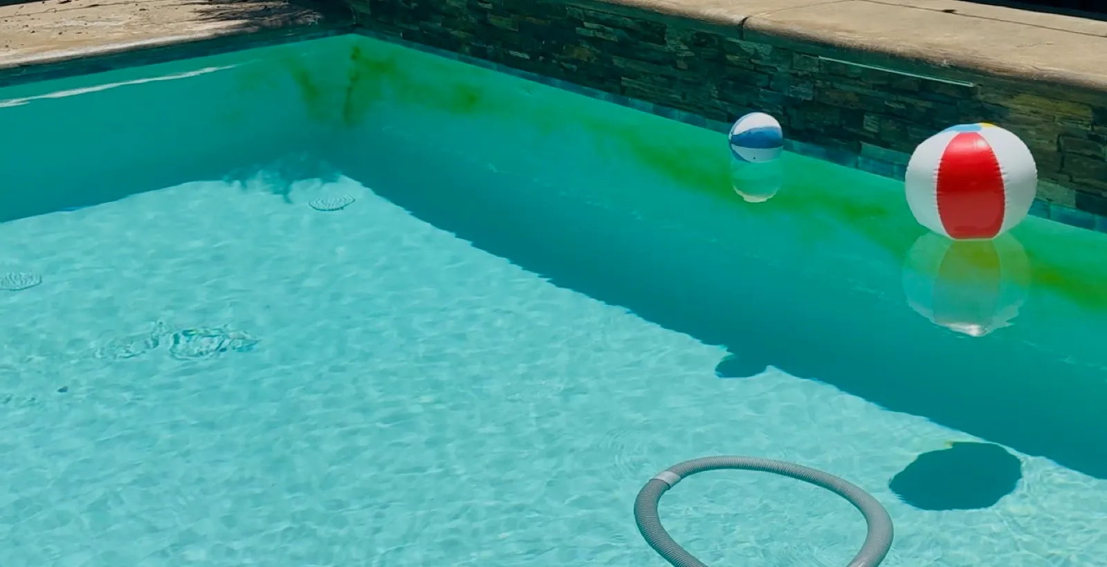 Rectangular pool with green algae streaks growing along the wall and beach balls floating in the water before professional pool algae shock treatment and vacuum service