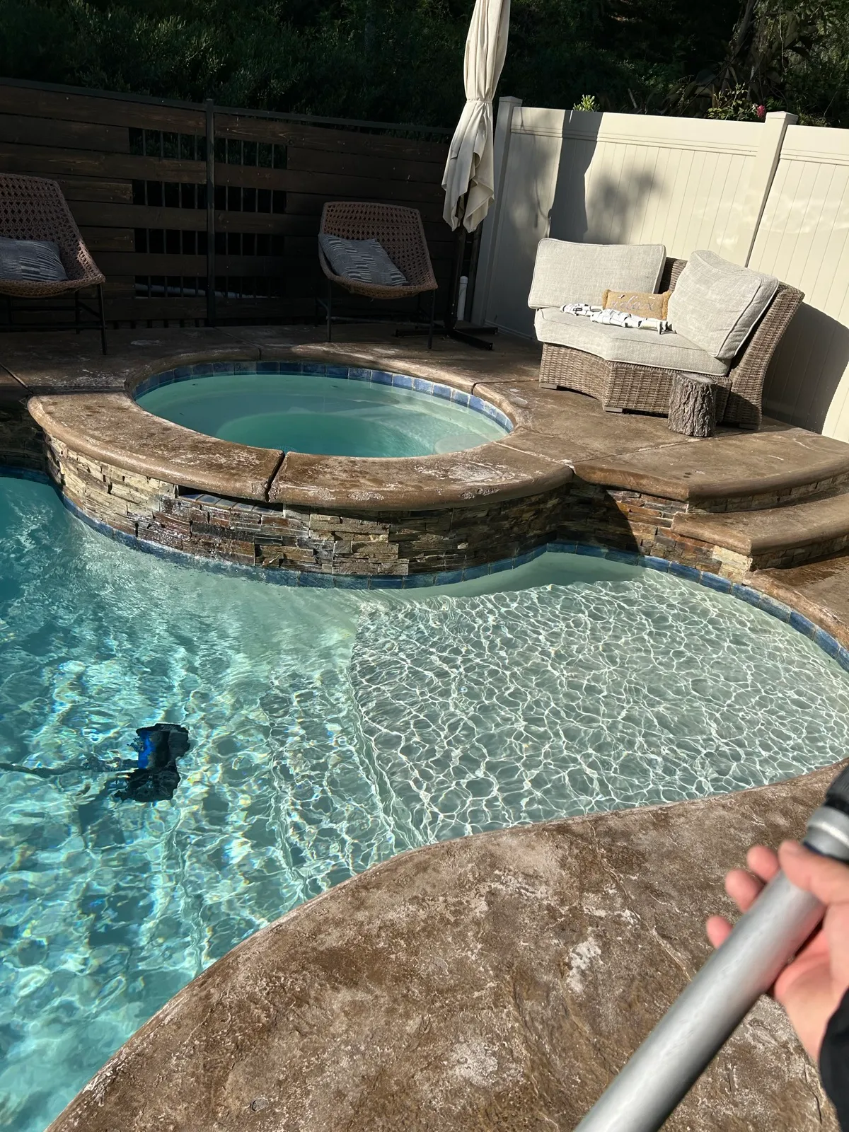 Pool service technician using a telescoping pole to clean a freeform backyard pool with attached round spa during a professional pool maintenance visit in Temescal Valley, CA